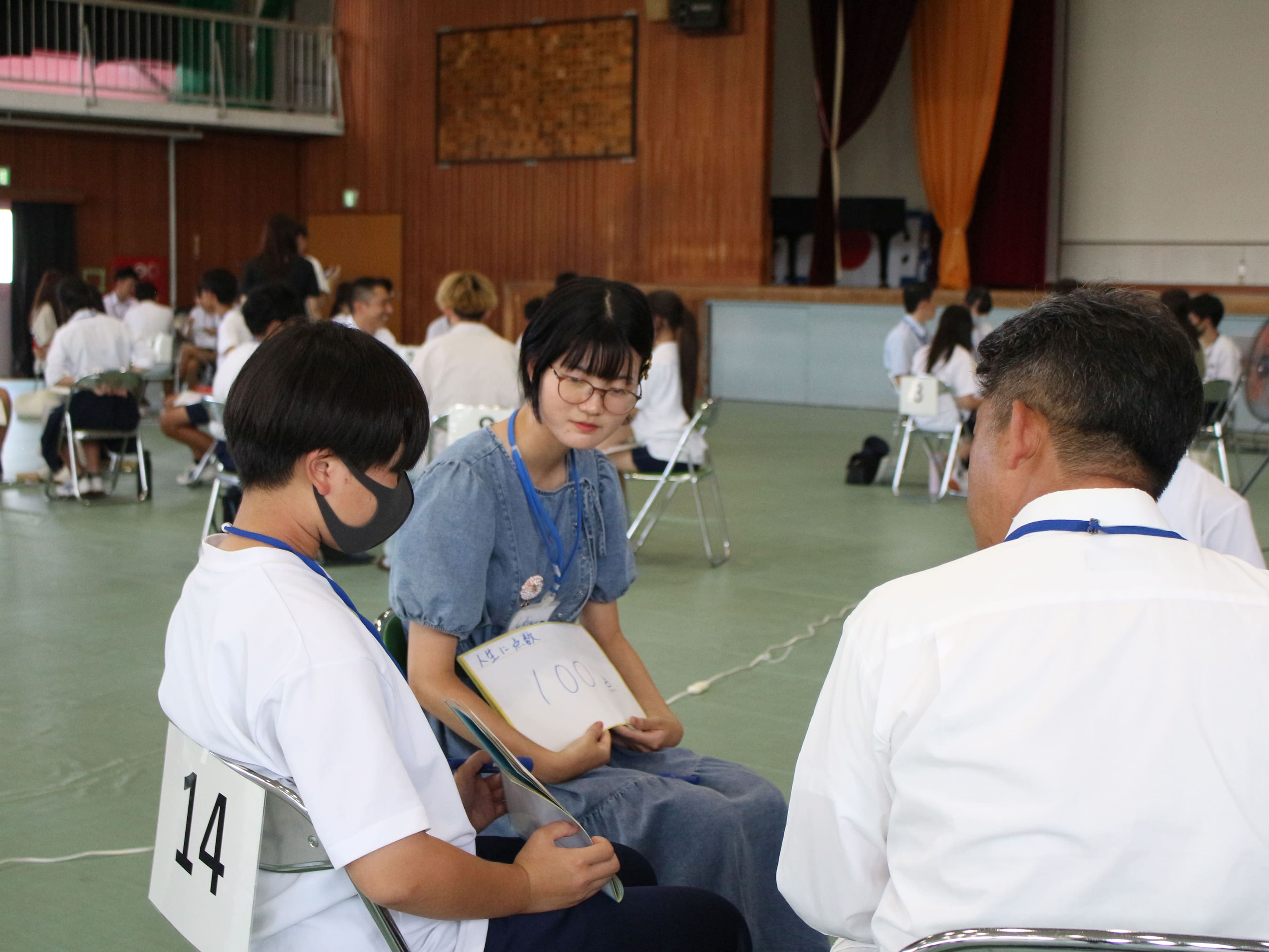 体育館の中でパイプ椅子に座り話を聞いている複数の中高生の様子